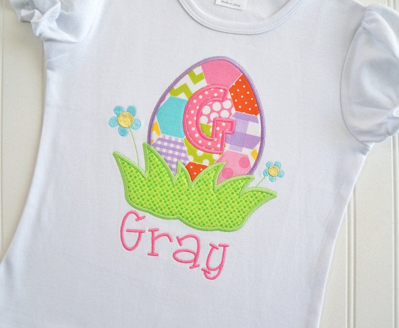 May include: White t-shirt with a colorful patchwork Easter egg design featuring the letter G and the name "Gray" embroidered in pink.