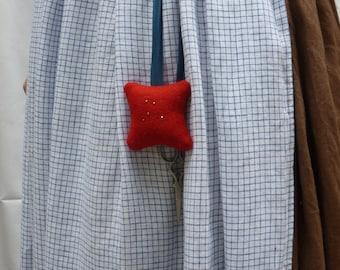 May include: A red pincushion with a blue ribbon and silver scissors hanging from it. The pincushion is attached to a white and blue gingham dress.