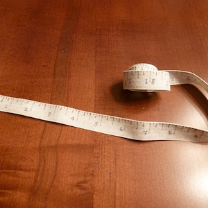 May include: A beige fabric measuring tape with black numbers, rolled up and laid out on a wooden surface. The tape measures from 1 to 9 inches.