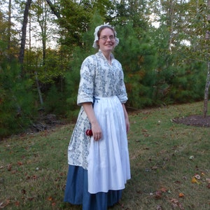 May include: A woman wearing a white and blue floral print dress with a white apron and a white bonnet. She is standing in a grassy area with trees in the background.