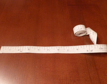May include: A white fabric measuring tape with black markings, showing inches and yards. The tape is laid out straight with the end rolled up.