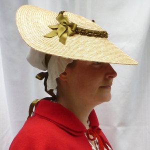 Puede incluir: Un sombrero de paja de ala ancha con una cinta y un lazo verdes. El sombrero lo lleva una persona con un gorro blanco y una chaqueta roja.