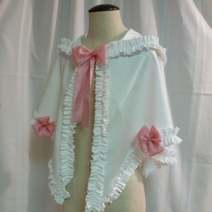May include: White hooded cape with ruffled trim and pink satin bows. The cape is made of a soft, lightweight fabric and has a relaxed fit.