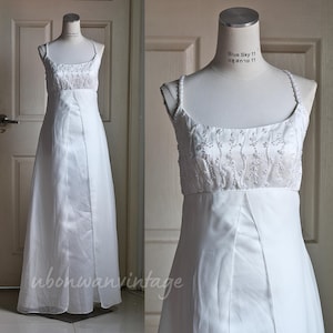 Puede incluir: Vestido de novia blanco sobre un maniquí. El corpiño está adornado con delicadas cuentas y bordados florales. El vestido tiene tirantes finos y una falda fluida. El vestido se exhibe frente a una puerta blanca.