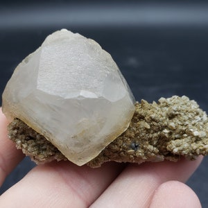 May include: A clear, translucent crystal with a pointed top, resting on a bed of brown and gray rock. The crystal is a natural specimen with a rough, textured surface.