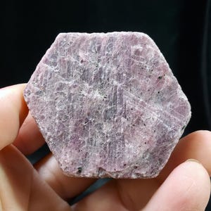 以下が含まれることがあります： 六角形のラフカットルビーの宝石。石は、より濃い紫と灰色の静脈を持つ、薄紫の色合いをしています。表面は質感があります。宝石は黒い背景に対して手で持たれています。
