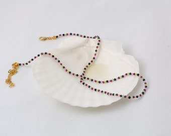 May include: Delicate necklace featuring small, colorful beads in pink, green, and dark blue. The necklace has a gold-colored chain and clasp. The jewelry is displayed on a white seashell against a white background.