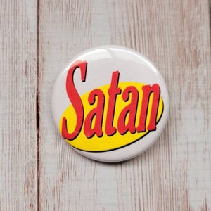 Puede incluir: Un botón blanco con la palabra "Satan" en rojo, texto estilizado, sobre un óvalo amarillo. El botón está sobre una superficie clara con vetas de madera.