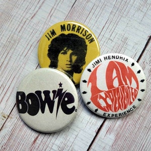以下が含まれることがあります： ミュージシャンの画像が描かれた丸いピンバッジが3つあります。最初のバッジは黄色で、白黒のジム・モリソンの画像が描かれています。2番目のバッジは白で、黒い文字で「ボウイ」と書いてあります。3番目のバッジは白で、赤い文字で「ジミ・ヘンドリックス・エクスペリエンス」と書いてあります。