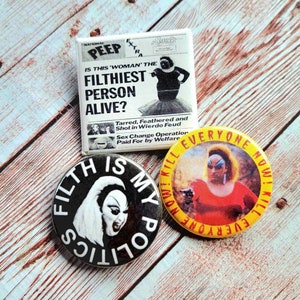 May include: A collection of three novelty buttons and a square pin. The square pin features a newspaper-style design with the headline "IS THIS 'WOMAN' THE FILTHIEST PERSON ALIVE?". The round buttons have the phrases "FILTH IS MY POLITICS" and "KILL EVERYONE NOW!".