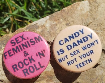 May include: Two circular badges with text. One is pink and reads "SEX, FEMINISM & ROCK 'N' ROLL" in black. The other is beige and says "CANDY IS DANDY BUT SEX WON'T ROT YOUR TEETH" in blue. They rest on a stone.