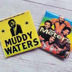 Peut inclure: Deux badges carrés. L'un est jaune avec une image en noir et blanc de Muddy Waters et le texte "Muddy Waters". L'autre badge présente un groupe d'hommes avec le texte "the Meters".