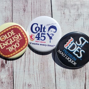 May include: Three circular pin badges featuring drinks branding. One is cream with red text for "OLDE ENGLISH 800". Another is white with blue text and a portrait for "Colt 45". The last is black with white and blue text for "ST DES Premium Malt Liquor".