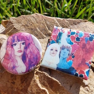 May include: Two round button pins with colorful illustrations. The first pin features a woman with long pink hair and a white dress. The second pin features three people with colorful backgrounds.