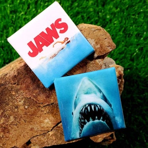 Peut inclure: Deux aimants carrés représentant le film "Jaws". L'un affiche le titre du film en rouge sur une scène océanique bleue. L'autre montre un gros plan de la gueule ouverte d'un requin avec des dents acérées.