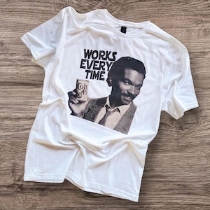 Könnte beinhalten: Weißes T-Shirt mit einem Schwarz-Weiß-Bild eines Mannes, der eine Colt 45-Dose hält. Der Text "WORKS EVERY TIME" ist über dem Bild aufgedruckt. Das T-Shirt hat einen Rundhalsausschnitt und ist aus weichem Material.