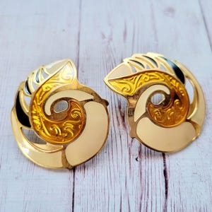 May include: A pair of gold-toned earrings with a spiral design. The earrings feature cream, gold, and black enamel accents. The design is reminiscent of a nautilus shell.