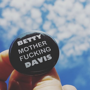 May include: A black button with white text that reads "BETTY MOTHER FUCKING DAVIS." The button is held up against a blue sky with white clouds.