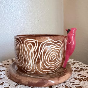 May include: A brown ceramic mug and matching saucer set featuring white hand-painted concentric circular patterns. The mug handle is sculpted in the shape of a bright pink cardinal bird with a black face mask, extending vertically from the side.