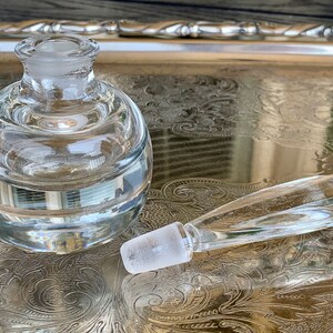 May include: A clear glass bottle with a round body and a separate glass stopper. The bottle has a textured surface and is sitting on a silver tray.