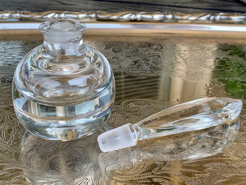 May include: A clear glass bottle with a round body and a separate glass stopper. The bottle has a textured surface and is sitting on a silver tray.
