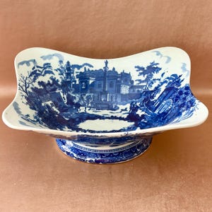 May include: A white and blue ceramic serving bowl with a pedestal base. The bowl features a blue landscape scene with buildings and trees. The pedestal base has a blue and white patterned design. The bowl has a unique, curved shape.