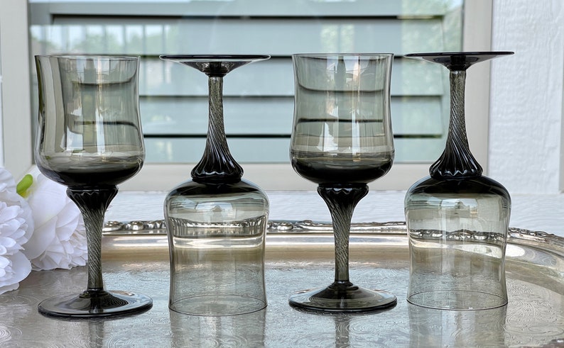 May include: Four clear glass wine glasses with a twisted stem and a dark gray tint. The glasses are arranged in a row on a silver tray.