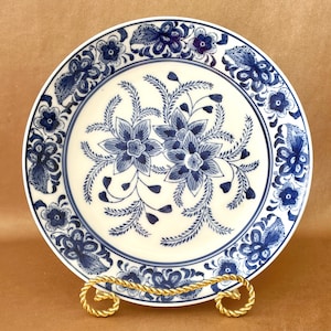 May include: A round, off-white ceramic plate with a blue floral design. The plate features a central cluster of flowers and a border of smaller floral patterns. The plate is displayed on a gold-colored metal stand.