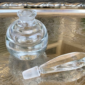May include: A clear glass bottle with a round body and a long, thin, clear glass stopper. The bottle is sitting on a silver tray with an ornate design.
