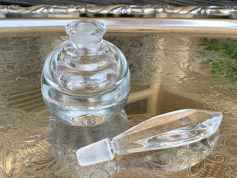 May include: A clear glass bottle with a round body and a long, thin, clear glass stopper. The bottle is sitting on a silver tray with an ornate design.