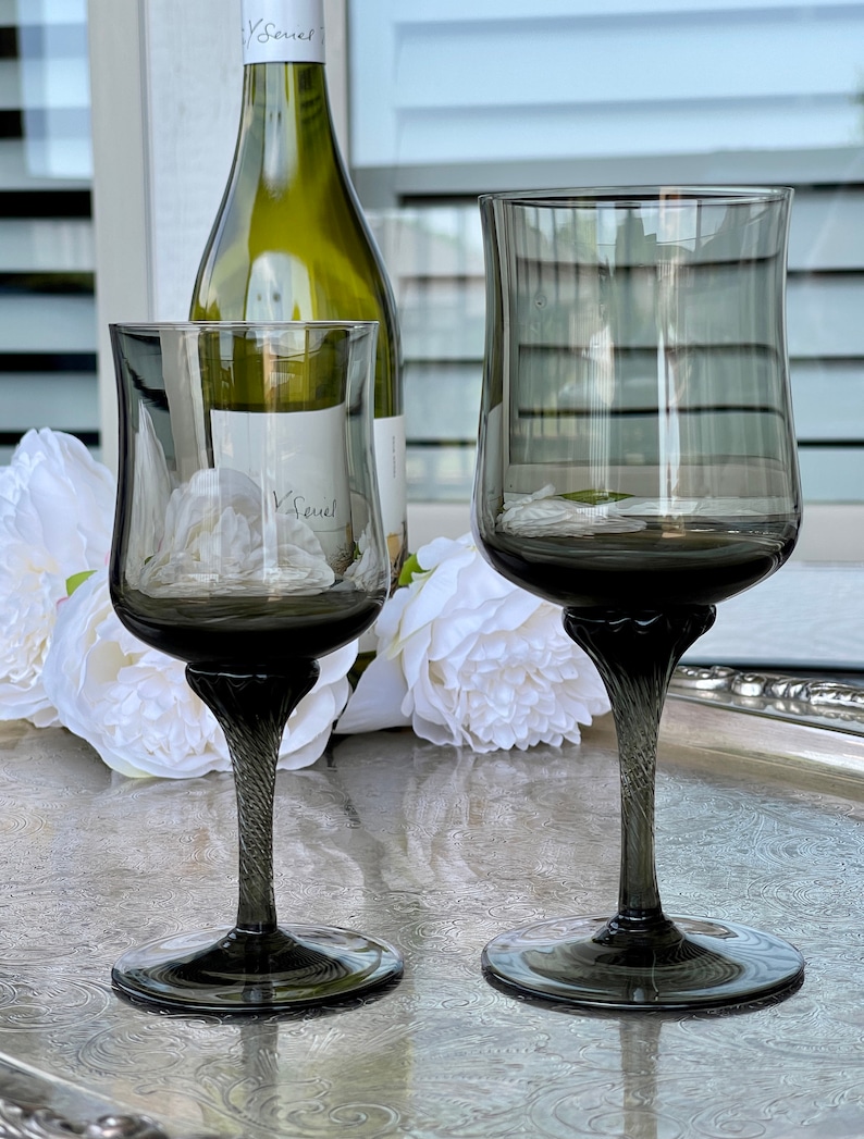 May include: Two dark gray glass wine glasses with a twisted stem on a silver tray with white flowers. The larger glass is taller and wider than the smaller glass.