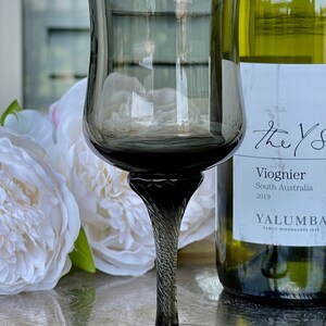 May include: A dark gray stemmed wine glass with a twisted stem sits on a silver surface next to a bottle of white wine. The bottle label reads "The Yarra Valley Viognier South Australia 2019 Yalumba Family Winemakers 1849."