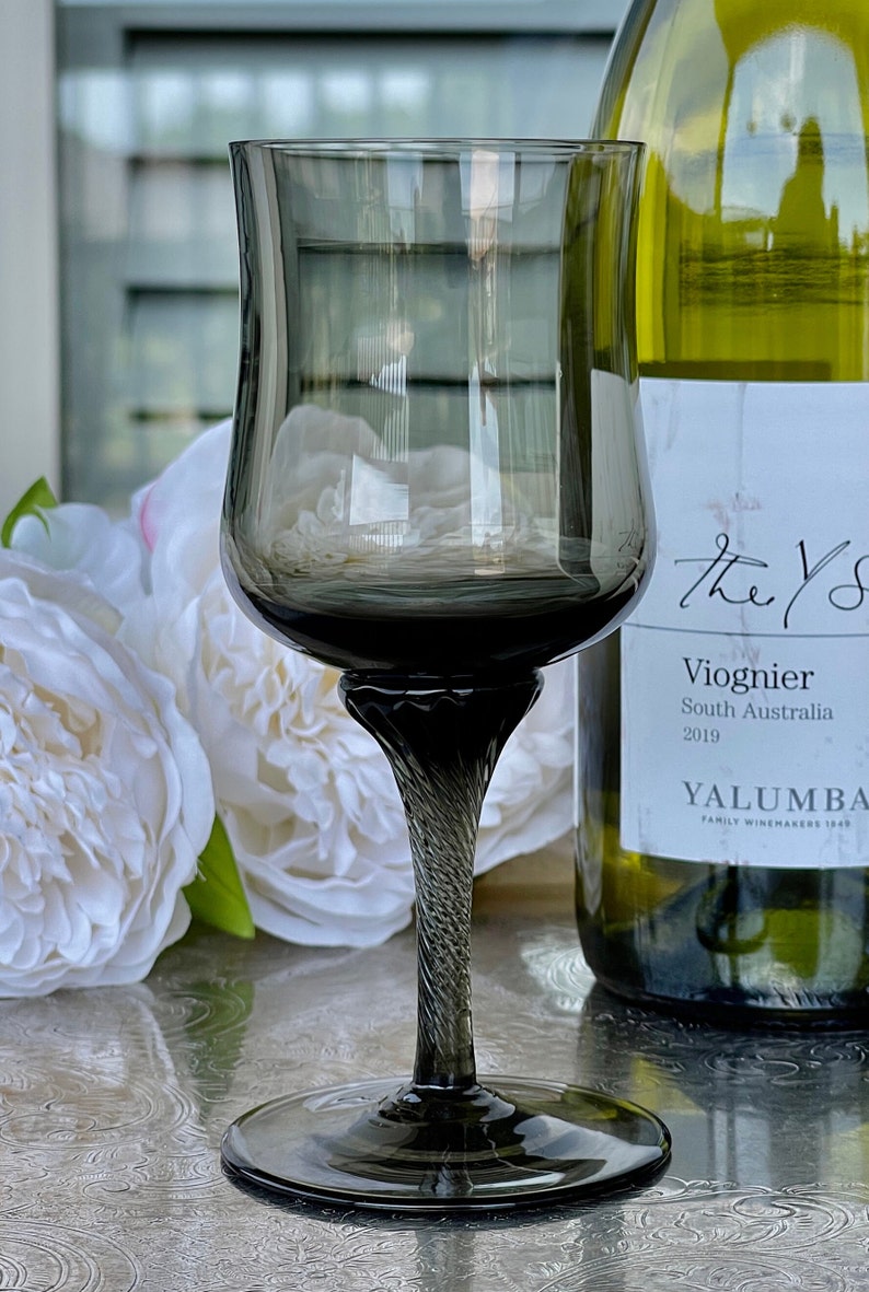 May include: A dark gray stemmed wine glass with a twisted stem sits on a silver surface next to a bottle of white wine. The bottle label reads "The Yarra Valley Viognier South Australia 2019 Yalumba Family Winemakers 1849."