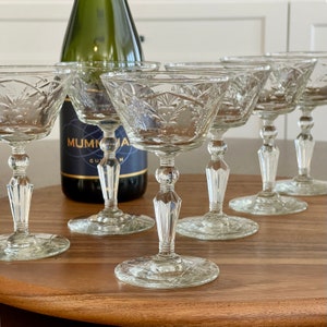 May include: Set of six clear glass champagne coupes with a cut glass design. The coupes have a round bowl and a tall, slender stem with a knob at the base.