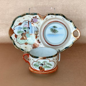 May include: A white porcelain teacup and saucer set with a divided tray. The tray features a Japanese-inspired design with a woman in a kimono and a landscape scene. The cup and saucer have a similar design with a woman in a kimono and a tree.