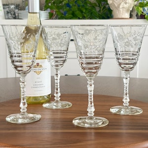 May include: Four clear, etched wine glasses with floral designs are arranged on a wooden surface. A bottle of white wine is positioned behind the glasses. The glasses have a detailed stem and a wide base, perfect for serving wine.