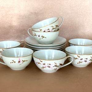 May include: Set of six white teacups and saucers with a delicate floral design in shades of pink and brown. The cups and saucers have a gold rim.