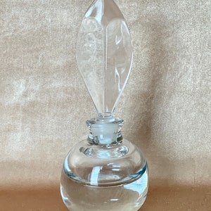 May include: A clear glass bottle with a round body and a pointed, leaf-shaped stopper. The bottle is filled with a clear liquid.