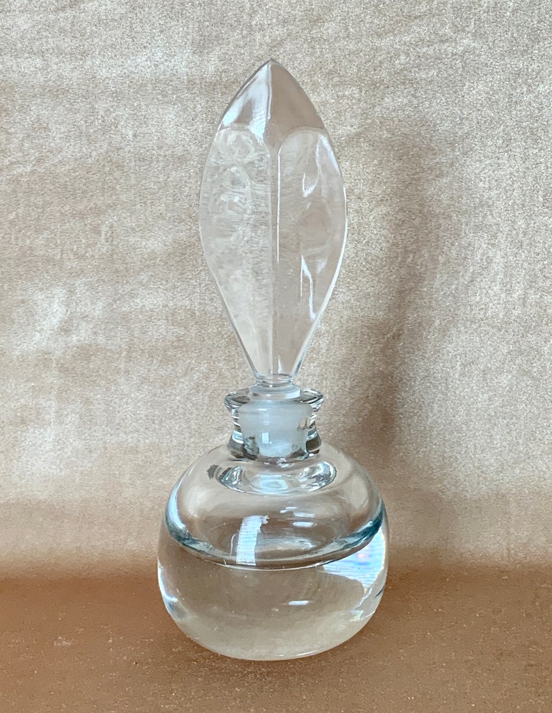 May include: A clear glass bottle with a round body and a pointed, leaf-shaped stopper. The bottle is filled with a clear liquid.