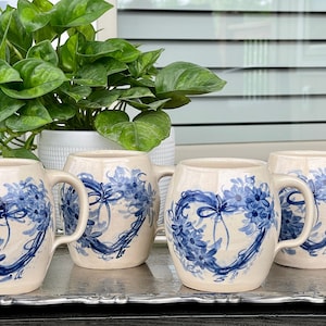 May include: Four ceramic mugs with blue and white floral designs. The mugs are heart-shaped and have a white background with blue floral wreaths.