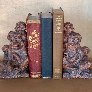 May include: A pair of bookends made of brown metal in the shape of monkeys. The monkeys are stacked on top of each other and are holding books. The books are titled "The Business Guide" and "The Secretary's Handbook".