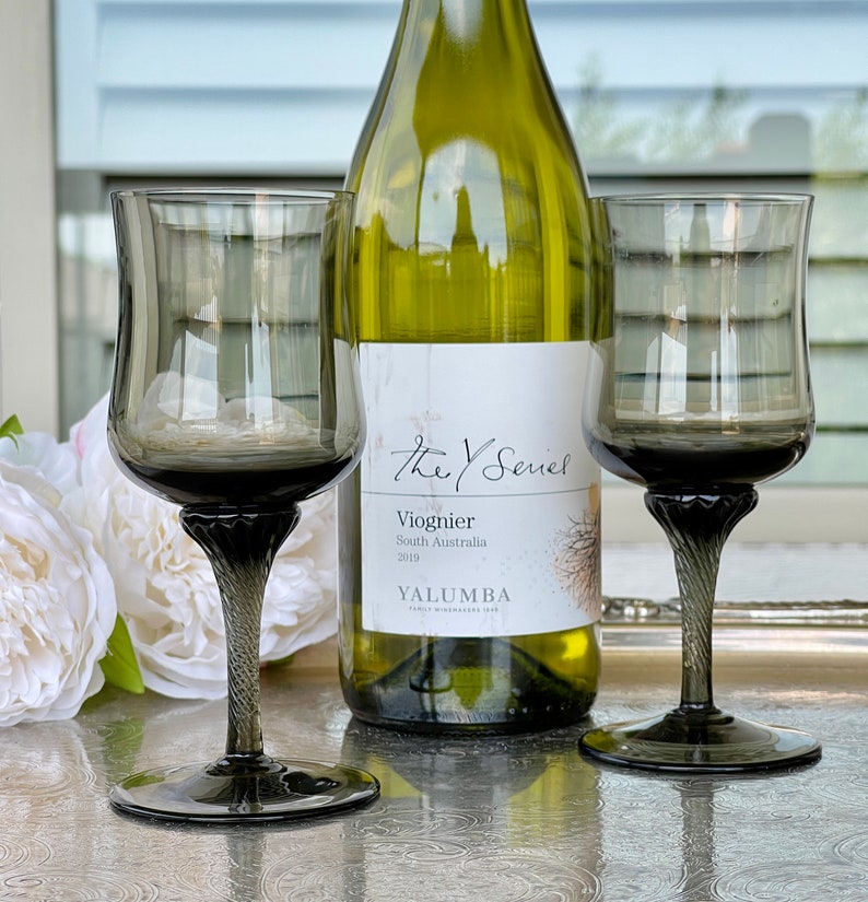 May include: Two dark gray stemmed wine glasses with a bottle of Y Series Viognier wine from Yalumba Family Winemakers. The bottle is green and has a white label with black text.