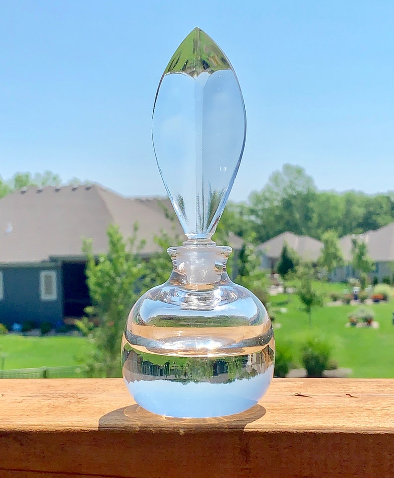 May include: A clear glass bottle with a pointed stopper. The bottle is round and has a textured surface. The bottle is sitting on a wooden surface.