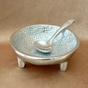 May include: A small, silver-colored bowl with a textured interior and three short legs. A matching spoon rests inside the bowl. The bowl and spoon are made of a shiny metal, likely aluminum or stainless steel.