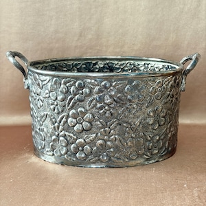 May include: An oval-shaped silver-toned metal basin featuring an embossed floral pattern across its exterior surface. The container includes two curved handles on opposite sides and a textured background behind the raised flower and leaf motifs.