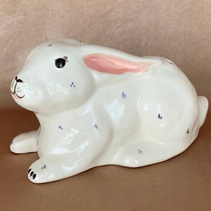 May include: A white ceramic bunny figurine with blue polka dots and a pink ear. The bunny is lying down with its head turned to the side.