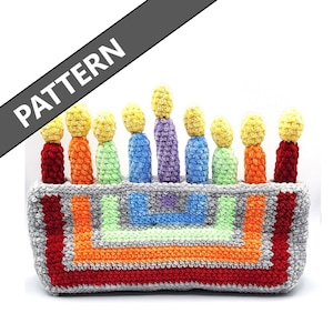 May include: A crocheted birthday cake with rainbow coloured candles. The cake is grey with orange, green, and red trim. The candles are made of yarn and have yellow tips.