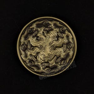 Puede incluir: Medallón circular de color bronce con un relieve detallado de un rostro con cabello ondeado. El diseño es intrincado, con patrones arremolinados y líneas finas. El medallón está sobre un fondo oscuro.