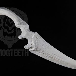 May include: A white, bone-shaped knife with a curved handle and a sharp blade. The handle has a textured appearance, and the blade is pointed. The word "SMOGTEETH" is printed in gray at the bottom of the image.