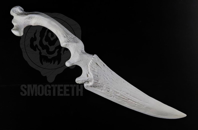 May include: A white, bone-shaped knife with a curved handle and a sharp blade. The handle has a textured appearance, and the blade is pointed. The word "SMOGTEETH" is printed in gray at the bottom of the image.